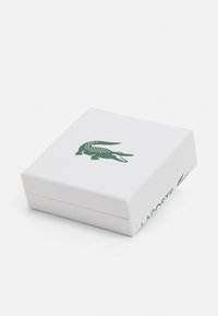 Caja rectangular blanca con un acabado texturizado, con un logo de cocodrilo verde en la tapa y "LACOSTE" impreso en el lateral.