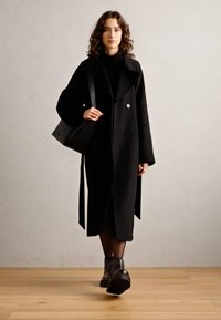 Lungo cappotto di lana nera con design doppiopetto, tasche laterali e cintura in vita. Il modello porta anche una borsa nera e indossa stivali chunky.
