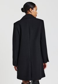 Donna con i capelli raccolti in un chignon, che indossa un lungo cappotto nero, vista di spalle su uno sfondo chiaro e uniforme.