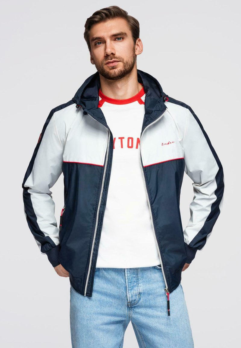 Giacca leggera da uomo con cappuccio, pannelli navy e bianchi, dettagli rossi, chiusura con zip e tasche laterali, abbinata a una t-shirt bianca.