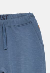 Blaue Zugbandhose mit elastischem Bund und Seitentaschen, strukturiertem Stoff und einem kleinen braunen Etikett an der rechten Seitennaht.