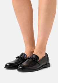 Svarta lädersneakers med kedjedetalj, bärs på bara fötter, visade framifrån på en enhetlig ljus bakgrund.