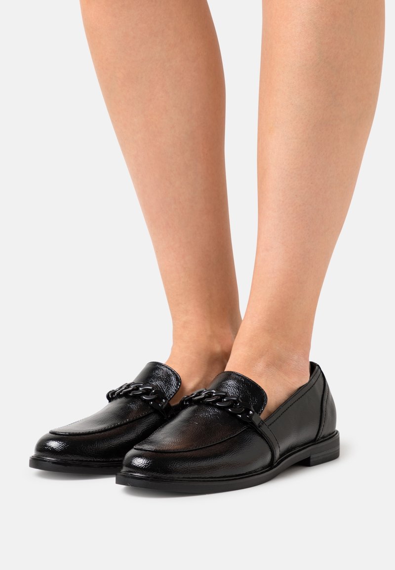 Svarta lädersneakers med kedjedetalj, bärs på bara fötter, visade framifrån på en enhetlig ljus bakgrund.