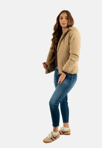 Giacca puffer beige con zip frontale, morbida fodera marrone e cappuccio, abbinata a jeans skinny blu e sneakers crema.