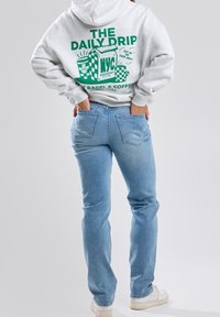 Grå huvtröja med grön grafisk text och illustrationer på baksidan, parat med ljusblå jeans och vita sneakers. Släta texturer, avslappnad passform.