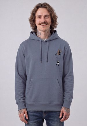 Lächelnder Mann mit welligem Haar, der einen grauen Hoodie mit einem Taschenmotiv mit kleinen Pandas und Essstäbchen trägt.