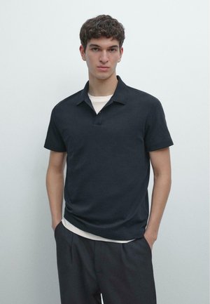 Giovane uomo con capelli corti e ricci, indossa una polo a maniche corte blu scuro sopra una T-shirt bianca, mani nelle tasche di pantaloni scuri, sfondo neutro.