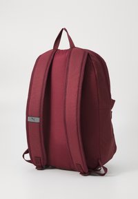 Burgundin reppu, jossa on pehmustetut hihnat, yläkahva, sileä pinta ja pieni harmaa logopatch sivulla. Vetoketjullinen pääosasto.