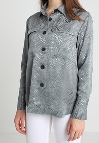 Grau langarmiges Shirt mit einem strukturierten, wellenförmigen Muster, ausgestattet mit einem Kragen, zwei Brusttaschen und schwarzen Knöpfen. Weiße Hose ist darunter sichtbar.