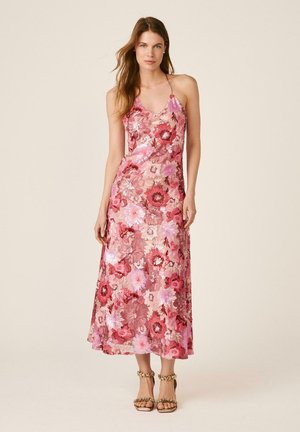 Mujer de pie con un vestido midi rosa con estampado floral y tirantes finos, y sandalias de tacón alto adornadas con cadenas doradas, sobre un fondo liso.