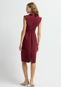 Robe ajustée bordeaux avec un col haut, des manches courtes, une taille ceinturée et une fente arrière. Texture lisse, conçue pour un ajustement sur mesure.