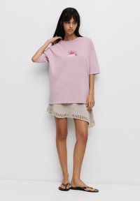Lyserød oversized t-shirt med et grafisk tryk i midten; båret over en beige strikskørt med en scalloperet kant og parret med sorte flip-flops.