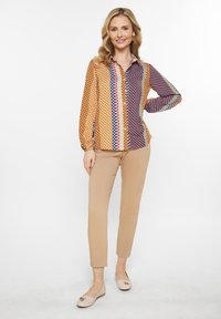 Multifarvet langærmet bluse med prikker og geometriske mønstre i orange, lilla og hvid, parret med beige tætsiddende bukser og lyse sko.