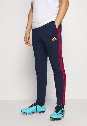 Mann trägt dunkelblaue Adidas-Trainingshose mit pinken Streifen und hellblaue Fußballschuhe, steht vor einem schlichten Hintergrund.