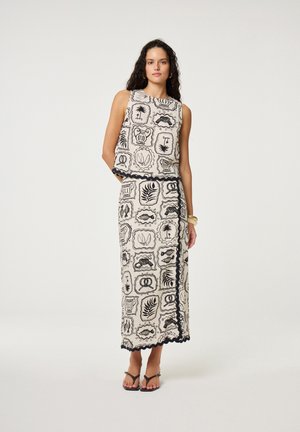 Vrouw met een mouwloze beige top en bijpassende lange rok met zwarte prints geïnspireerd op de natuur en met gegolfde randen, staand tegen een effen achtergrond.