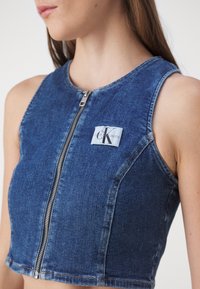 Vrouw met een mouwloos blauw denim crop top met rits en een Calvin Klein Jeans logo patch op de borst.