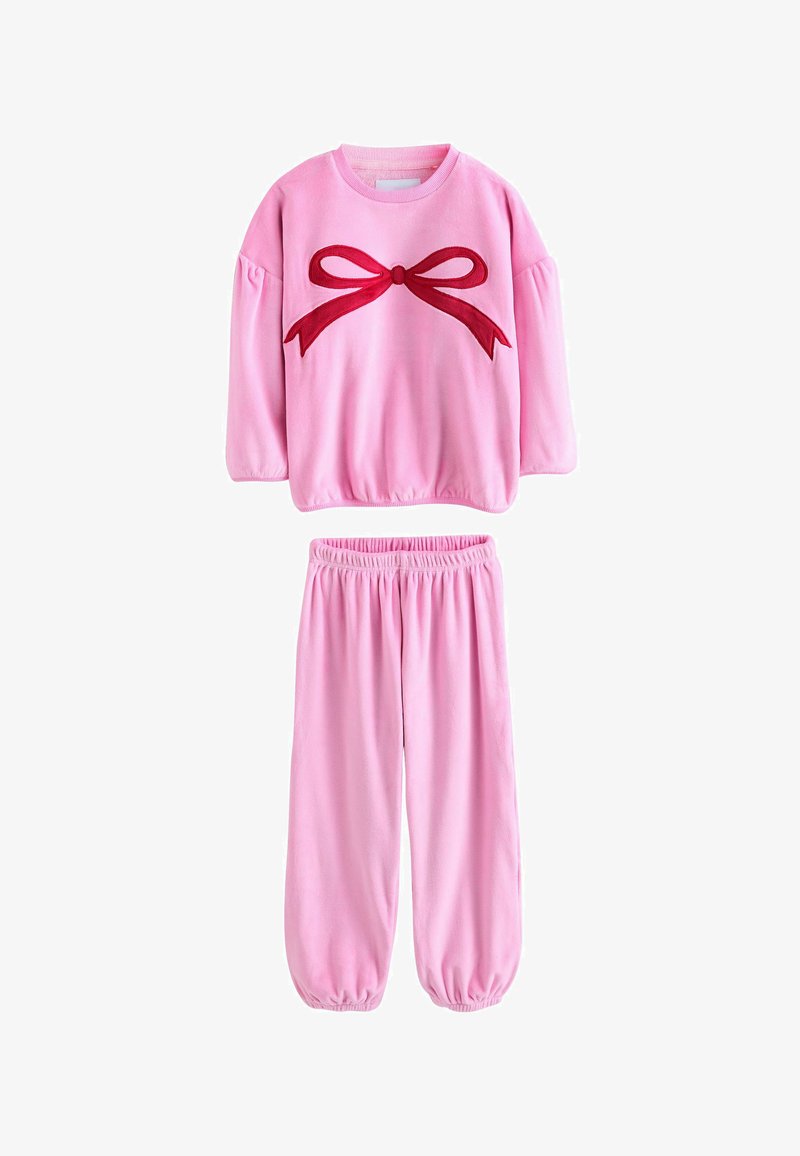 Tuta in velour rosa con collo a giro, maniche a palloncino e applicazione di un fiocco rosso sulla parte superiore. Vita elasticizzata sui pantaloni affusolati.