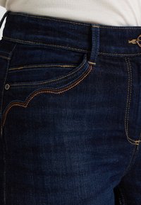 Pantalones vaqueros de mezclilla azul oscuro con cintura alta, costuras en naranja y marrón, y bolsillos delanteros con una ligera curva en la costura.