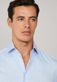 Mlad muškarac s kratkom tamnom kosom koji nosi svijetloplavu košulju s gumbima, gleda u stranu naslonjen na jednostavnu pozadinu.