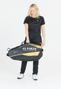 Musta ja kultainen FZ Forza urheilulaukku, tyylikäs muotoilu, jossa on teksturoitu kuvio ja logo, jota kantaa henkilö mustassa urheiluasussa.
