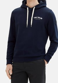 Marinblå hoodie med ribbat mönster, snören och en vit logotyp på bröstet. Har en rund patch på vänster ärm.