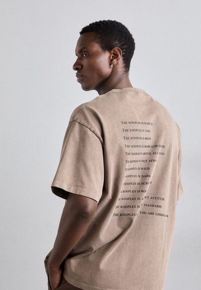 Camiseta de algodón marrón con mangas cortas, que presenta texto impreso en negro en la parte posterior. El texto incluye varias frases sobre "The Kooples".