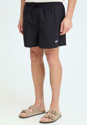 Männliche Beine, die schwarze Shorts mit Kordelzug und Logo am linken Bein sowie beigefarbene Sandalen mit zwei Riemen tragen, stehen vor einem einfarbigen hellen Hintergrund.