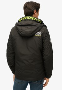 Zwarte gewatteerde jas met capuchon, voorzien van een gestructureerde stof, felgroen logo op de kraag en mouw, en een slank, getailleerd ontwerp.