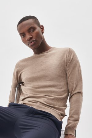 Suéter beige de manga larga con cuello redondo, hecho de tela texturizada. El modelo está sentado en una silla de metal, vistiendo pantalones azul marino.