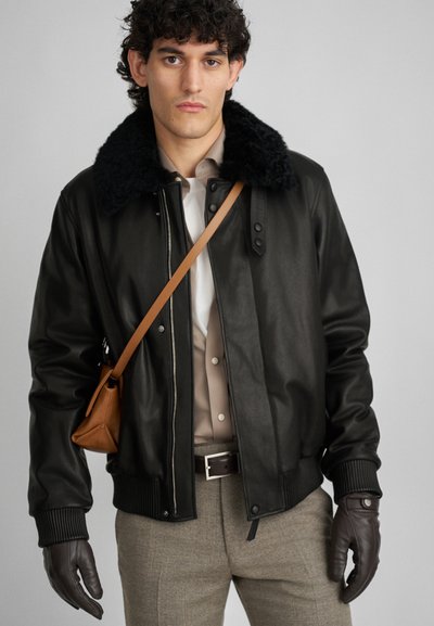 Veste bomber en cuir noir avec un col en fourrure, des poignets côtelés et une fermeture éclair à l'avant. Associée à une chemise beige et à un pantalon gris. Sac bandoulière marron.