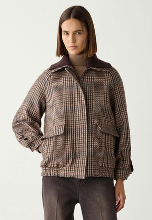 Frau trägt eine braune karierte Jacke über einem braunen Rollkragenpullover und steht vor einem schlichten hellen Hintergrund.