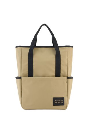 Beige Tragetasche mit schwarzen Griffen und Verzierungen, Fronttasche, Seitentaschen und einem schwarzen "Studio Berlin"-Etikett in der rechten unteren Ecke.