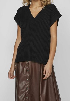 Femme portant un pull noir côtelé à manches courtes en V et une jupe en cuir marron à boutons, debout devant un fond uni.