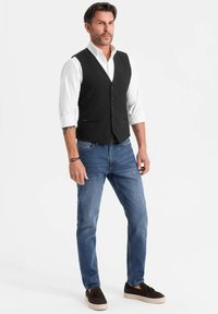 Uomo in piedi che indossa una camicia bianca con maniche arrotolate, un gilet nero, jeans blu e mocassini marroni con nappe su uno sfondo bianco.
