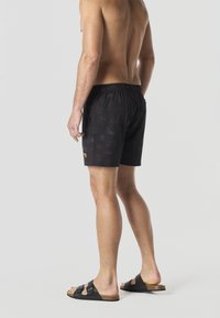 Uomo senza maglietta che indossa pantaloncini da bagno neri con motivo e sandali neri a ciabatta, in piedi davanti a uno sfondo chiaro uniforme, visto di schiena.