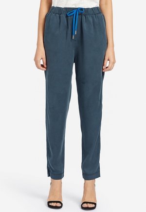 Vrouw draagt donkerblauwe broek met trekkoord en zwarte open sandalen, staand tegen een witte achtergrond.