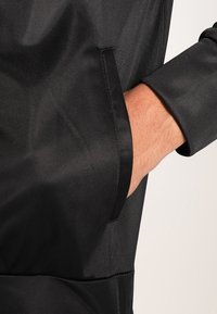 Svart jacka med ett slätt, texturerat tyg. Har en sidficka och raka ärmar. Förstärkta sömmar är synliga.