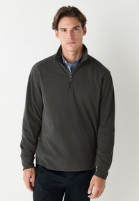 Tumši zaļš villaina džemperis ar rāvējslēdzēju, augstu apkakli, garām piedurknēm un viendabīgu faktūru. Ir ceturkšņa garuma rāvējslēdzējs un ribotas aproces.