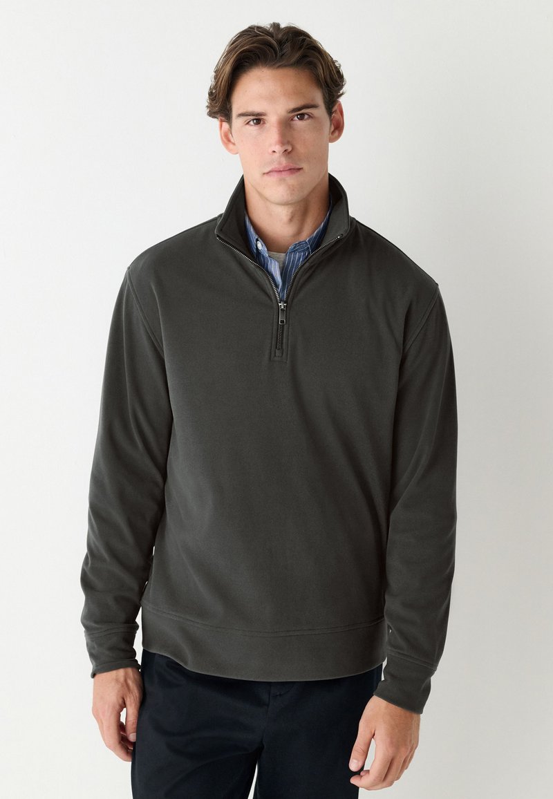 Tumši zaļš villaina džemperis ar rāvējslēdzēju, augstu apkakli, garām piedurknēm un viendabīgu faktūru. Ir ceturkšņa garuma rāvējslēdzējs un ribotas aproces.