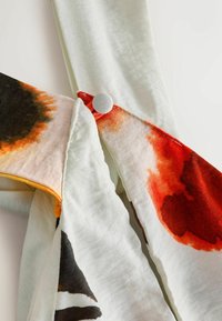 Tissu blanc avec de grandes impressions florales abstraites rouges, orange et noires et un bouton blanc fixant deux sections ensemble.