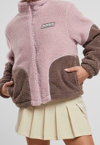 Chaqueta de forro polar con un cuerpo rosa y bolsillos marrones, con cuello alto, cierre de cremallera y detalles en blanco. Ideal para mantener el calor y la comodidad.