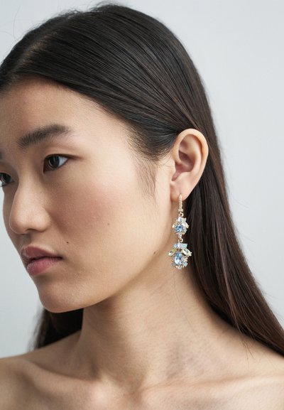 Zlatne viseće naušnice s plavim i prozirnim dragim kamenjem, s cvjetnim dizajnom. Dragulji su postavljeni u delikatnu metalnu strukturu s teksturom.