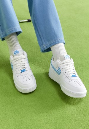 Osoba nosząca białe buty Nike Air Force 1 z niebieskim kratowanym logo, białe skarpetki i jasnoniebieskie spodnie stojąca na zielonym dywanie.