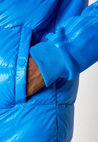 Mão no bolso de uma jaqueta acolchoada azul brilhante usada sobre uma jaqueta de treino azul com punho canelado e detalhe listrado na manga.