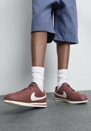Kind met witte sokken en maroon Nike sneakers met witte swoosh, staand op een grijze grond tegen een effen muur.