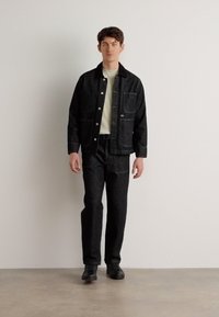 Giacca di denim nero e pantaloni con cuciture visibili, tasche frontali e chiusura con bottoni. Indossato con un maglione chiaro e scarpe nere.