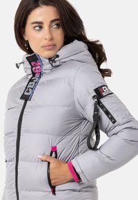 Hellgraue Pufferjacke mit Kapuze, mit schwarzen und pinken Akzenten, Reißverschlusstaschen und einem Patch am Ärmel. Glatte, gesteppten Textur.