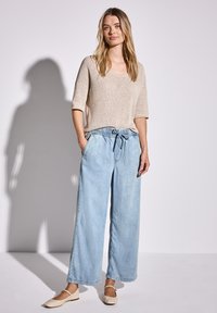 Beige Strickpullover mit kurzen Ärmeln, hellblaue weit geschnittene Jeans mit Bindegürtel und beigefarbene Ballerinas, vor einem hellgrauen Hintergrund.