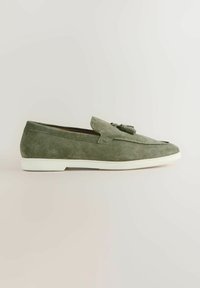 Mocassin en daim vert olive avec pompons et semelle en caoutchouc blanche, présenté de profil sur un fond neutre.