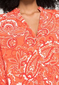 Rote Bluse mit floralem Muster, die einen V-Ausschnitt hat und aus einem weichen, leichten Stoff mit aufwändigen weißen und dunkler roten Designs gefertigt ist.